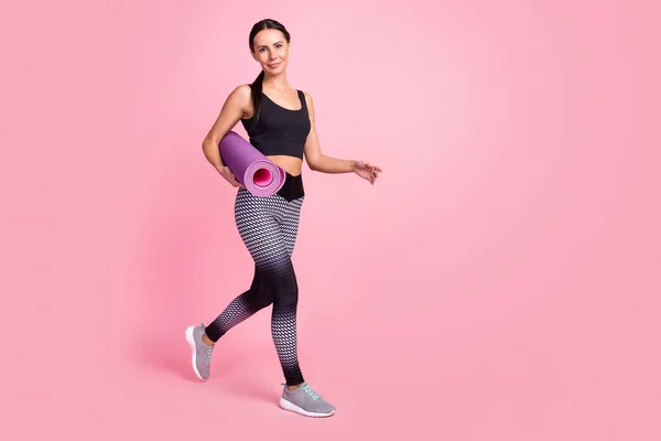 Foto de mujer joven linda positiva vestida traje deportivo caminando sonriente sosteniendo estera enrollada espacio vacío aislado color rosa fondo —  Fotos de Stock