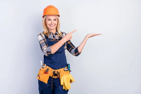 Photo of pretty positive mature handywoman workwear hardhat smiling pointing holding arm empty space isolated grey color background — Stock Photo, Image