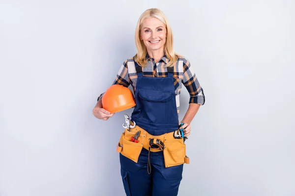 Photo of cheerful petty mature handywoman workwear smiling holding hardhat isolated grey color background — Stock Photo, Image