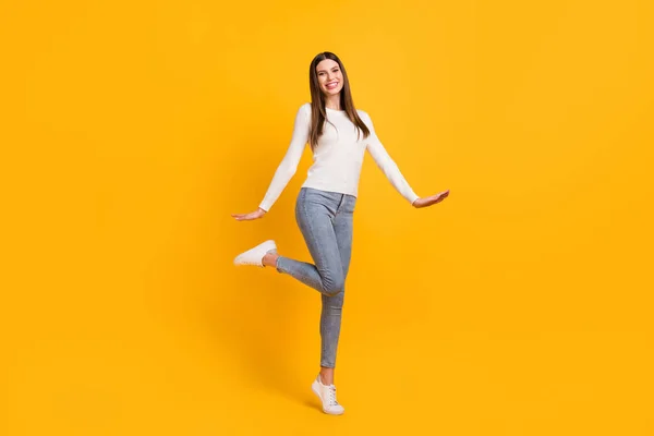 Foto de tamaño completo de pie chica morena optimista usar suéter blanco jeans zapatillas aisladas en el fondo de color amarillo —  Fotos de Stock