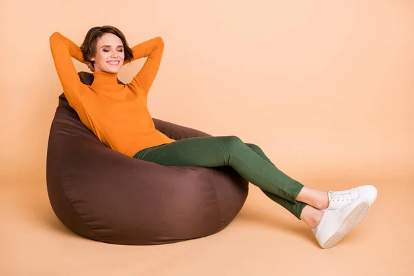 Foto de tamaño completo de joven bonita chica positiva feliz disfrutando de tiempo libre en sillón aislado sobre fondo de color beige —  Fotos de Stock