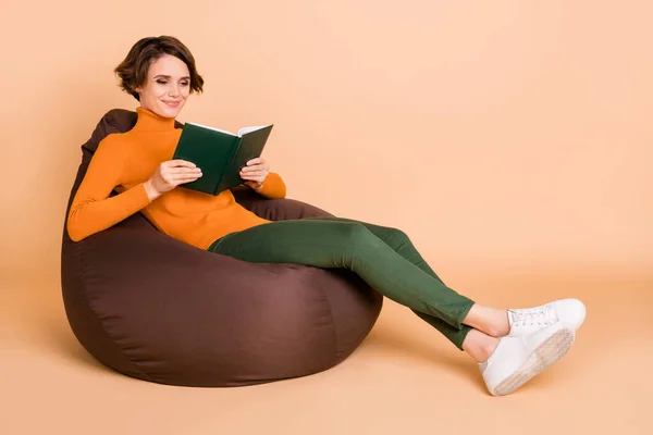 Teljes hosszúságú fotó bájos fiatal lány narancs garbó ülő bab táska olvasás könyv elszigetelt bézs színű háttér — Stock Fotó