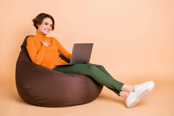 Foto seukuran penuh gadis cantik yang tersenyum gembira duduk di kursi berlengan menahan laptop yang terisolasi dengan latar belakang warna krem — Stok Foto