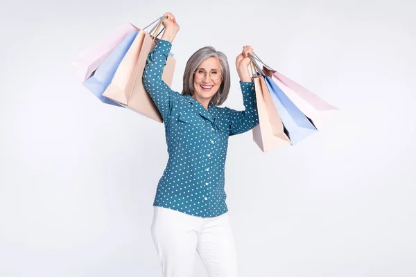 Fénykép a boldog pozitív idős nő emelje bevásárló táskák mosoly viselje szemüveg fehér nadrág elszigetelt szürke színű háttér — Stock Fotó