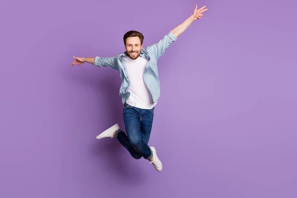 Фото возбужденного смешного молодого человека носить джинсовую рубашку прыгая высоко поднимая руки стороны стороны фиолетового цвета фона — стоковое фото