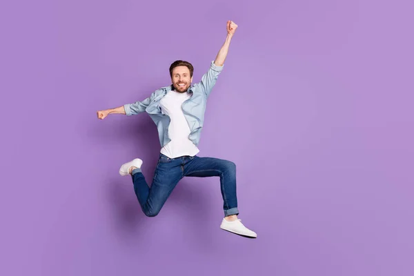 Фото очаровательного молодого человека носить джинсы рубашку прыгая высоко поднимающийся кулак улыбаясь пустое пространство изолированный фиолетовый цвет фона — стоковое фото