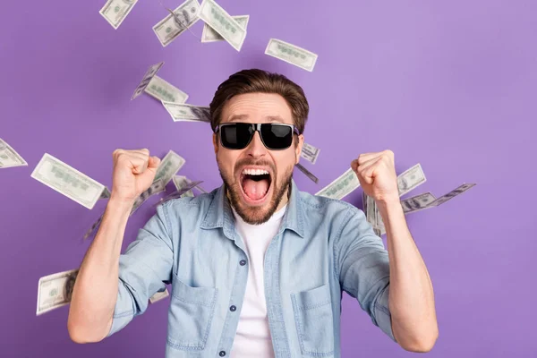 Foto de chico joven afortunado impresionado vestido de mezclilla gafas oscuras dinero volando puños ascendentes aislado color púrpura fondo — Foto de Stock