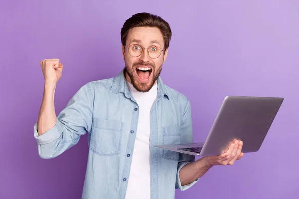 Φωτογραφία από σοκαρισμένος χαρούμενος χαρούμενος νεαρός άνδρας raise fist hold υπολογιστή νικητής απομονώνονται σε μωβ φόντο χρώμα — Φωτογραφία Αρχείου
