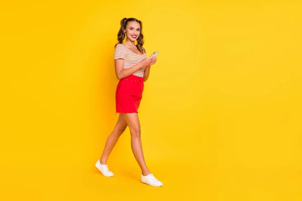 Foto de menina encantadora andar segurar telefone usar camisa listrada mini saia tênis isolado cor amarela fundo — Fotografia de Stock