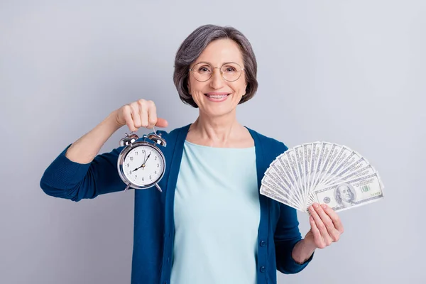 Foto ritratto di allegra donna d'affari che mostra sveglia mantenendo le banconote in dollari isolate su sfondo di colore grigio — Foto Stock