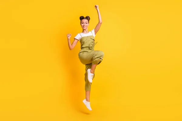 Foto de tamaño completo de la mujer morena optimista salto manos puños desgaste brillante camiseta general zapatillas aisladas en el fondo de color amarillo — Foto de Stock