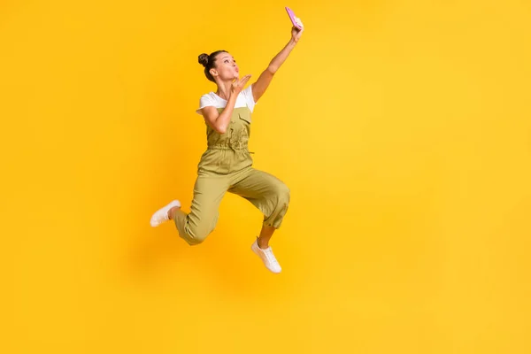 Tam boy vücut ölçülerinde güzel bir kızın atlama görüntüsü selfie çekerken parlak sarı arka planda izole edilmiş hava öpücüğü gönderiyor. — Stok fotoğraf