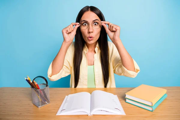 Foto de chocado jovem mulher usar óculos sentar mesa estudo exame livro inesperado isolado no fundo de cor azul — Fotografia de Stock