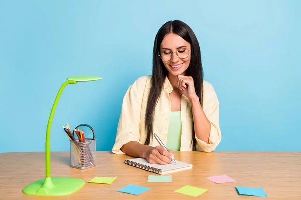 Портрет привлекательной веселой девушки руки на подбородке написания сидя за рабочим столом изолированы на синем фоне цвета — стоковое фото