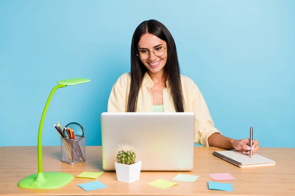 Foto de alegre jovem morena olhar laptop sentar mesa trabalho wtite nota isolada no fundo cor azul pastel — Fotografia de Stock