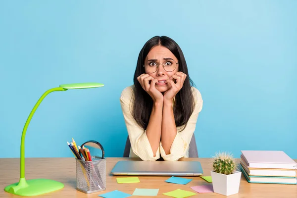 Retrato de senhora aterrorizada sentado atrás de desktop mordendo unha dedo olhando câmera isolada no fundo de cor azul — Fotografia de Stock