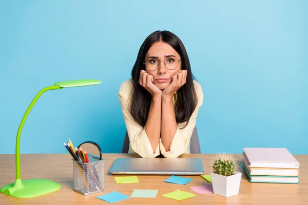 Foto de mujer joven cansada infeliz cogida de la mano cara sentarse trabajador de escritorio aislado sobre fondo de color azul pastel —  Fotos de Stock