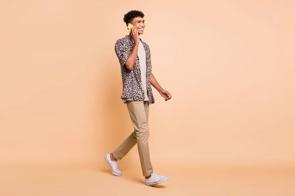 Full size profiel foto van brunet optimistische man gaan praten telefoon dragen moderne shirt broek sneakers geïsoleerd op beige achtergrond — Stockfoto