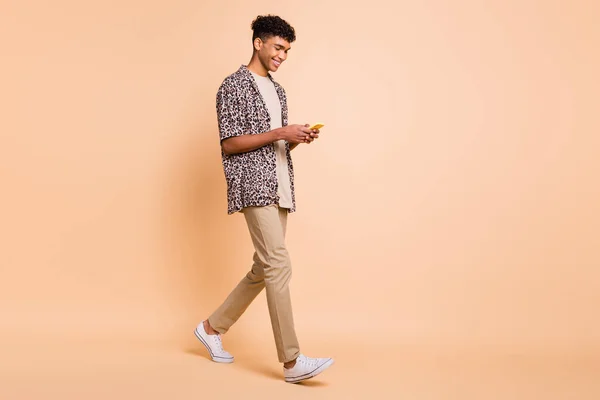 Full size profiel foto van brunet optimistische man gaan kijken telefoon dragen moderne shirt broek sneakers geïsoleerd op beige achtergrond — Stockfoto