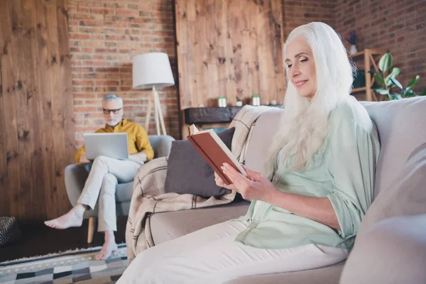 Fénykép a nyugdíjas ősz hajú nyugdíjas család ülni kanapé olvasni könyv használata laptop beltéri házban lakás — Stock Fotó