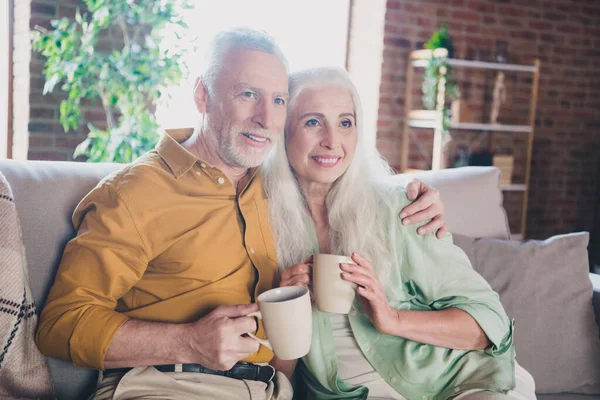 Retrato de atractiva pareja afectuosa alegre de pelo gris sentada en el sofá abrazando la bebida en casa casa loft de ladrillo casa interior — Foto de Stock
