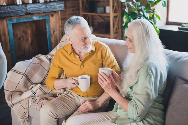 Foto de positivo encantador hombre y mujer ancianos buen humor sentarse sofá beber té en el interior de la casa de casa —  Fotos de Stock