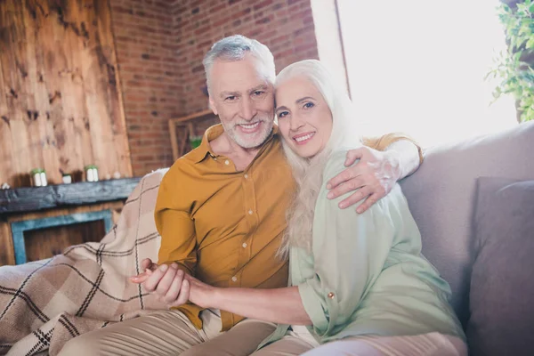 Foto de feliz casal de idosos positivo marido e mulher sentar sofá sorriso abraço segurar as mãos dentro de casa fim de semana — Fotografia de Stock