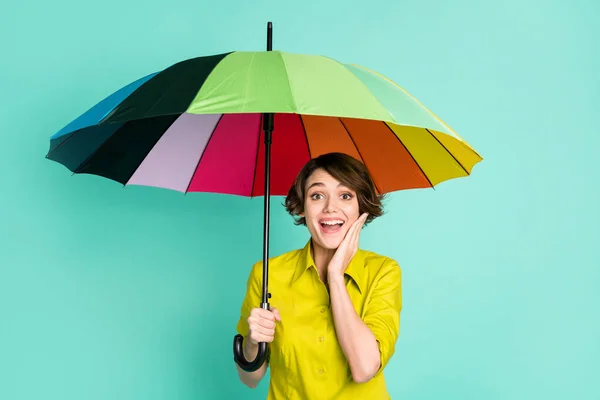 Фотопортрет изумленной улыбающейся девушки, касающейся щеки, держащей зонтик изолированным на ярком бирюзовом фоне — стоковое фото