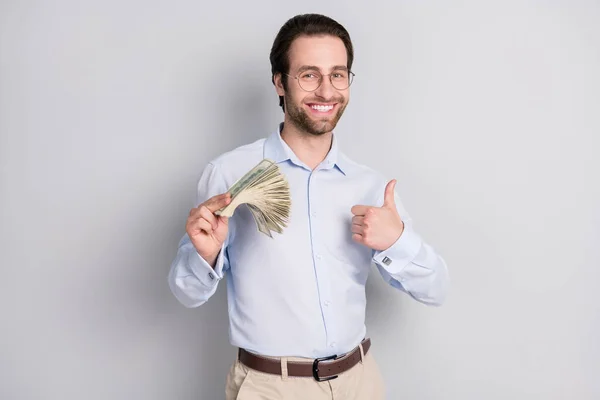 Retrato de hombre alegre de moda guapo sosteniendo en dinero manojo mostrando pulgar aislado sobre fondo de color pastel gris — Foto de Stock