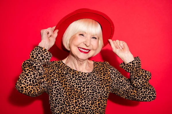 Фото ошеломляющей счастливой старой женщины носить красивую шляпу улыбку хорошее настроение изолированы на красном фоне цвета — стоковое фото