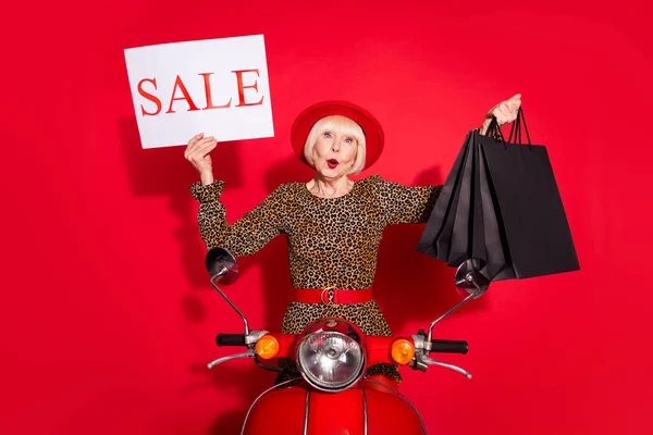 Foto di donna anziana cavalcare moto tenere banner vendita tenere borse della spesa nero venerdì isolato su sfondo di colore rosso — Foto Stock