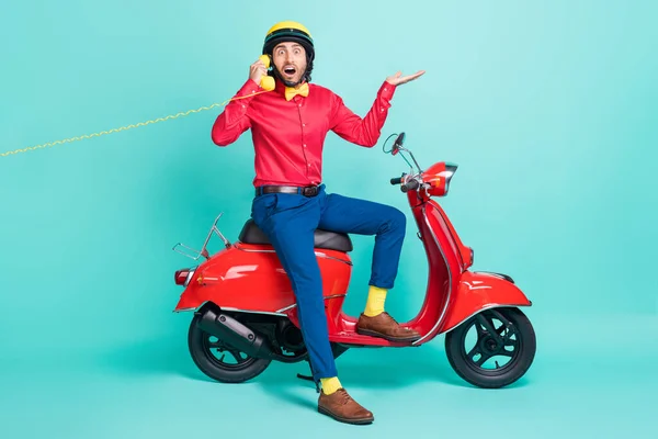 Foto em tamanho completo de jovem chocado homem surpreso sentar ciclomotor falando no telefone isolado no fundo cor turquesa — Fotografia de Stock