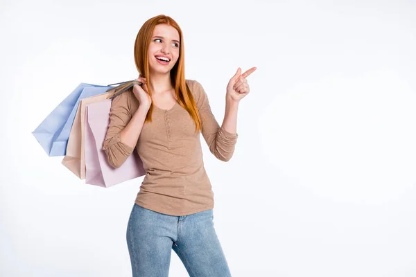 Foto de encantadora mujer pelirroja impresionada camisa beige sosteniendo bolsas de tienda apuntando con el dedo espacio vacío aislado color blanco fondo — Foto de Stock