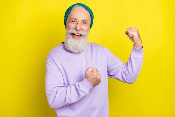 Foto de dulce afortunado hombre maduro vestido púrpura jersey headwear sonriente levantamiento puños aislado color amarillo fondo — Foto de Stock