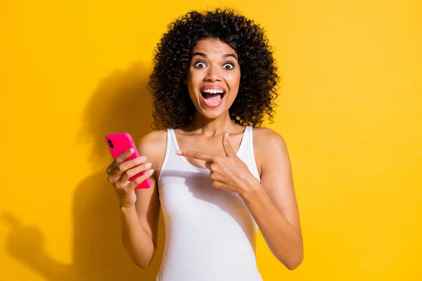 Foto de la piel oscura asombrada morena lady point teléfono usar singlet blanco aislado en el fondo de color amarillo vibrante — Foto de Stock