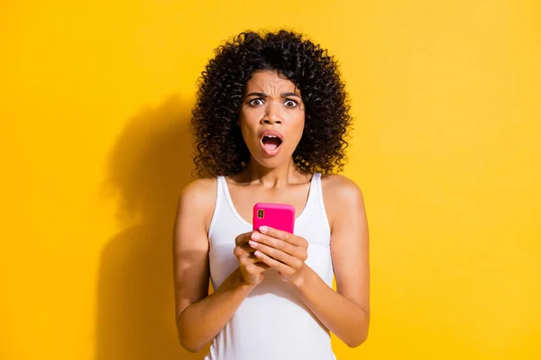 Foto de morena asombrada asustada mujer afro americana sostiene teléfono usar camiseta blanca aislada sobre fondo de color amarillo — Foto de Stock