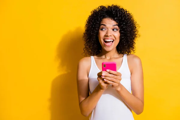 Photo of dark skin brown haired woman look empty space hold phone wear white singlet izolowane na żółtym tle koloru — Zdjęcie stockowe