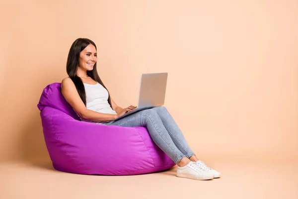 Foto potret seluruh tubuh wanita dengan laptop duduk di kursi beanbag terisolasi di latar belakang berwarna pastel beige — Stok Foto