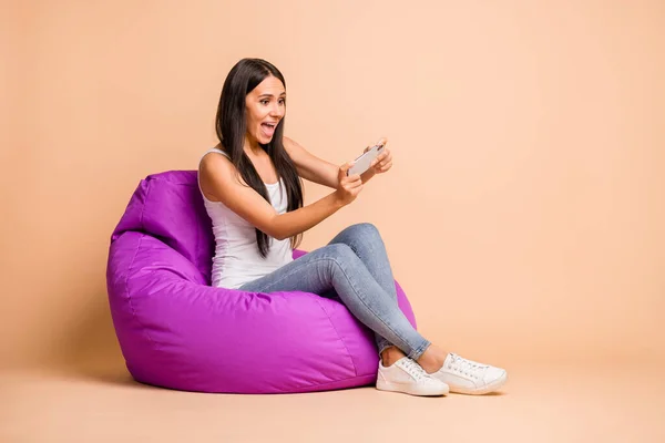 Foto porträtt helkroppsbild av kvinna spelar spel som håller telefonen vertikalt sitter isolerad på pastell beige färgad bakgrund — Stockfoto