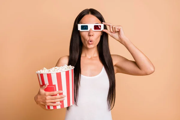 Photo portrait of impressed woman touching 3d glasses holding big popcorn bag isolated on pastel beige colored background — Stock Photo, Image