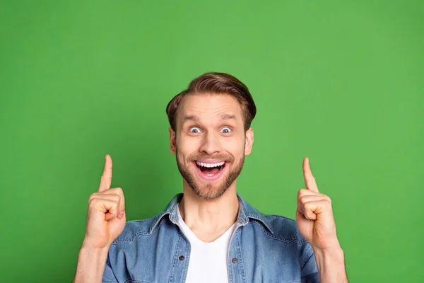 Фотопортрет изумленного парня, указывающего пальцем на пустое пространство, рекомендующий улыбаться изолированно на ярком зеленом фоне — стоковое фото
