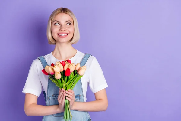 Foto van mooie optimistische korte kapsel blonde dame houden bloemen kijken lege ruimte dragen witte t-shirt geïsoleerd op violette achtergrond — Stockfoto