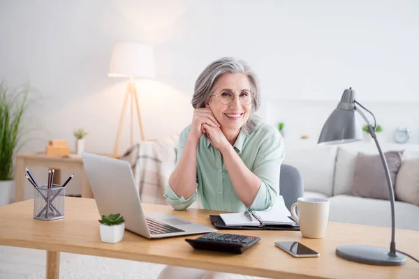 Foto de senhora muito positiva sentar atrás das mãos da mesa na bochecha sorriso olhar câmera tem bom humor dentro de casa — Fotografia de Stock