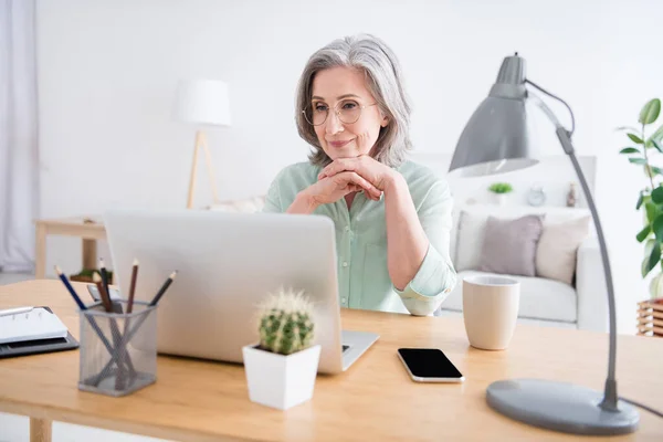 Portret van minded gerichte vrouw zit achter desktop armen onder kin kijken geïnteresseerd laptop binnen — Stockfoto