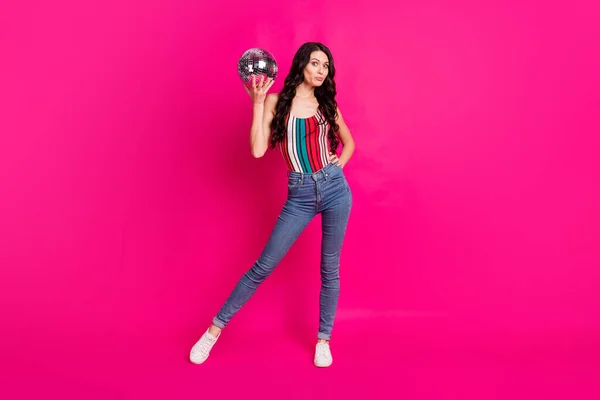 Foto de tamaño completo de la mujer morena optimista sostener disco bola usar top jeans zapatillas aisladas sobre fondo de color rosa vivo —  Fotos de Stock