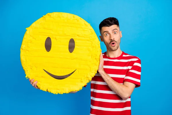 Foto de asombrado joven sorprendido sostiene papel grande sonrisa de piñata cara emoji aislado sobre fondo de color azul —  Fotos de Stock