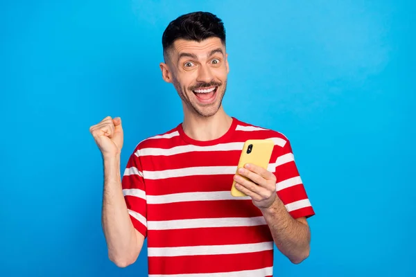 Foto de triunfante joven feliz levantar el puño sostener el teléfono como celebrar aislado en el fondo de color azul pastel — Foto de Stock