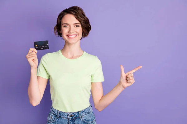 Porträtt av härlig glad flicka som håller i händerna bankkort visar åt sidan kopiera utrymme annons isolerad över violett färg bakgrund — Stockfoto