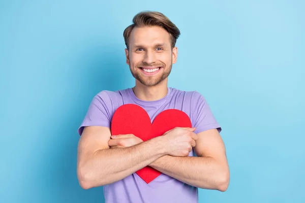 Foto retrato de un joven abrazando el corazón rojo usando una camiseta púrpura aislada sobre fondo de color azul pastel — Foto de Stock