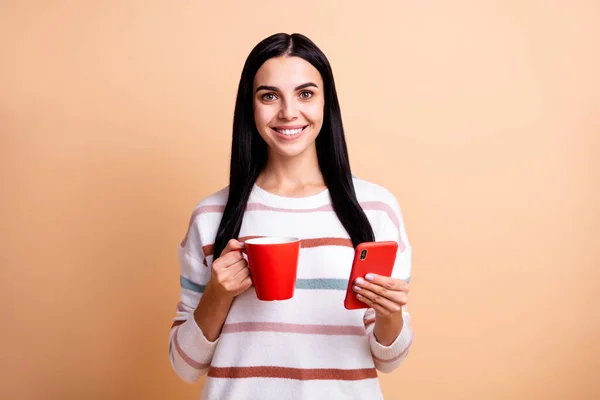 Fénykép a fiatal vonzó lány boldog pozitív mosoly élvezze reggeli csésze kávé használata mobiltelefon elszigetelt bézs színű háttér — Stock Fotó